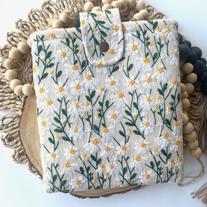 K&ouml;nnte beinhalten: Eine wei&szlig;e Stofftasche mit einem Blumenmuster aus wei&szlig;en G&auml;nsebl&uuml;mchen und gr&uuml;nen Stielen. Die Tasche hat einen Klappverschluss mit einem braunen Knopf.