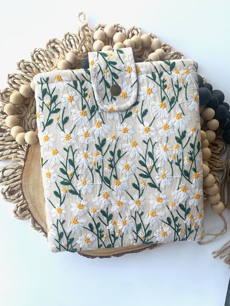 K&ouml;nnte beinhalten: Eine wei&szlig;e Stofftasche mit einem Blumenmuster aus wei&szlig;en G&auml;nsebl&uuml;mchen und gr&uuml;nen Stielen. Die Tasche hat einen Klappverschluss mit einem braunen Knopf.