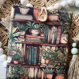 May include: A black fabric book sleeve with a bookshelf design featuring books, plants, and pots. The design is in shades of green, brown, and cream. A button is attached to the sleeve.