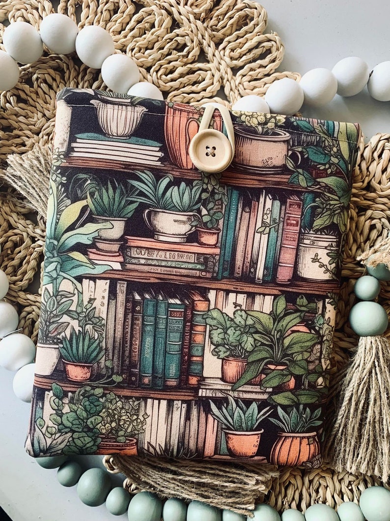 May include: A black fabric book sleeve with a bookshelf design featuring books, plants, and pots. The design is in shades of green, brown, and cream. A button is attached to the sleeve.