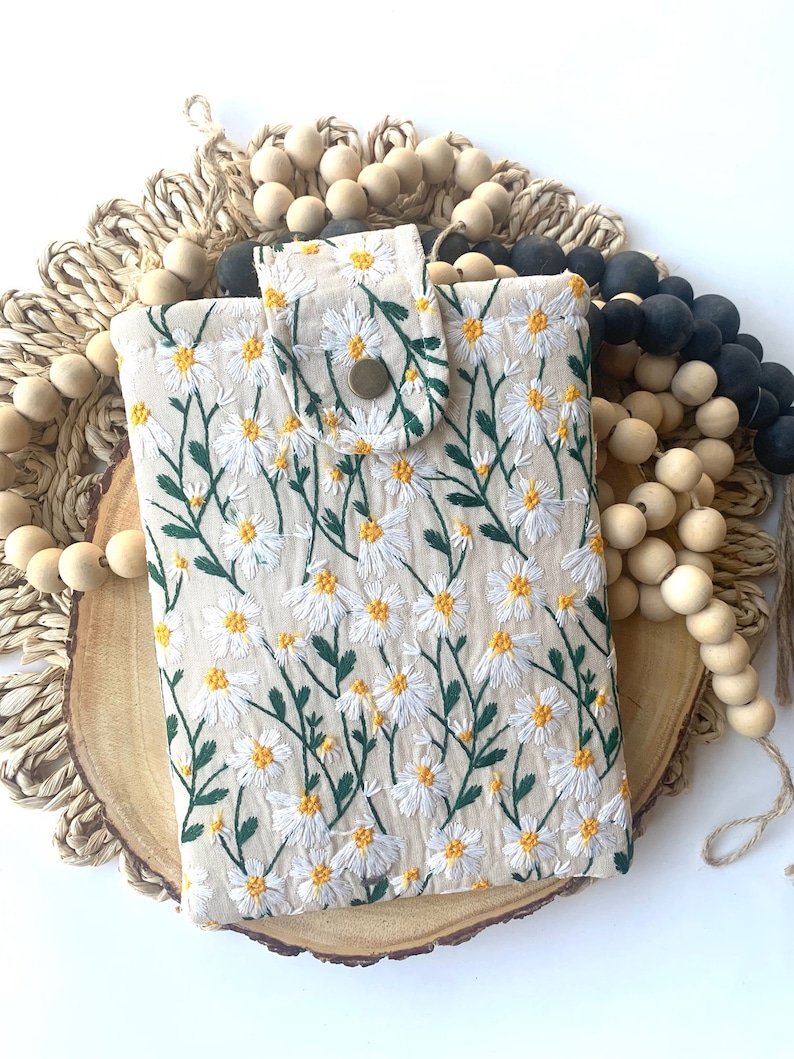 K&ouml;nnte beinhalten: Eine beige Handyh&uuml;lle mit einem gestickten Muster aus wei&szlig;en G&auml;nsebl&uuml;mchen. Die H&uuml;lle hat eine Klappe mit einem Knopfverschluss.