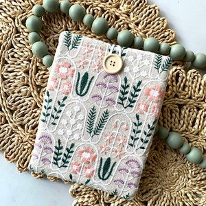 May include: Beige embroidered tablet case with a floral design in pink, white, and purple. The case has a button closure and is set on a woven placemat with a strand of green wooden beads.