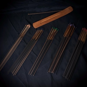May include: A wooden incense burner with a long, narrow groove and a single incense stick resting on it, accompanied by five distinct bundles of incense sticks featuring varied colored tips including blue, green, purple, and red.