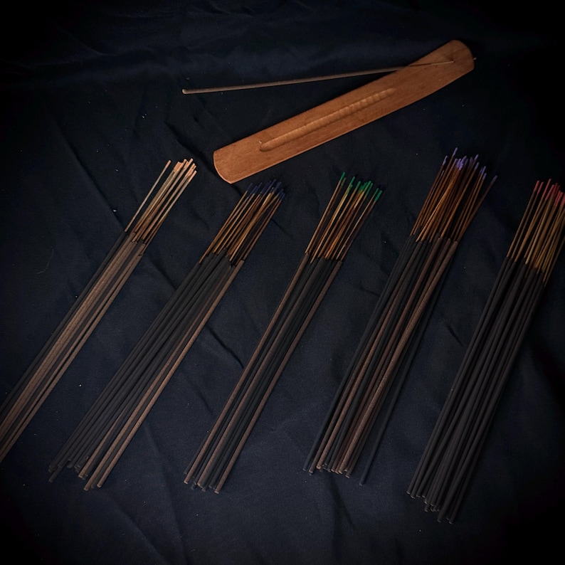 May include: A wooden incense burner with a long, narrow groove and a single incense stick resting on it, accompanied by five distinct bundles of incense sticks featuring varied colored tips including blue, green, purple, and red.
