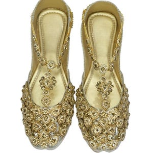 May include: A pair of gold embellished flat shoes with a t-strap design. The shoes are covered in small, sparkling rhinestones.