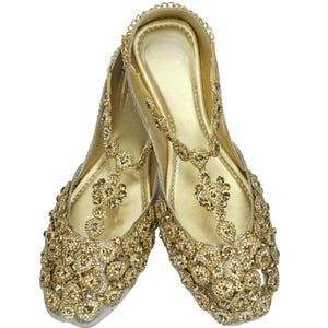 May include: A pair of gold embellished flat shoes with intricate detailing. The shoes have a pointed toe and a delicate ankle strap.