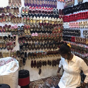 K&ouml;nnte beinhalten: Eine Wand voller bunter bestickter Schuhe in verschiedenen Stilen und Farben. Die Schuhe sind in Reihen an einer wei&szlig;en Wand mit einem wei&szlig;en Gittermuster angeordnet. Eine Person steht vor der Wand und betrachtet die Schuhe.