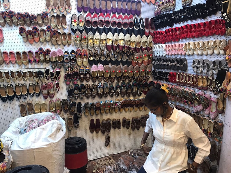 K&ouml;nnte beinhalten: Eine Wand voller bunter bestickter Schuhe in verschiedenen Stilen und Farben. Die Schuhe sind in Reihen an einer wei&szlig;en Wand mit einem wei&szlig;en Gittermuster angeordnet. Eine Person steht vor der Wand und betrachtet die Schuhe.