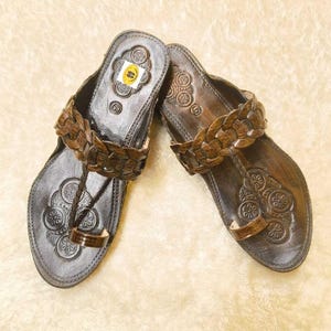 May include: A pair of dark brown leather sandals featuring a braided strap across the top and a toe loop. The footbed is embossed with circular floral patterns and includes a small yellow sticker with the number 6 near the heel area.