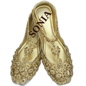 May include: A pair of gold embellished flat shoes with a t-strap design. The shoes are decorated with rhinestones and have a gold chain detail. The word "SONIA" is written on the insole of one shoe.