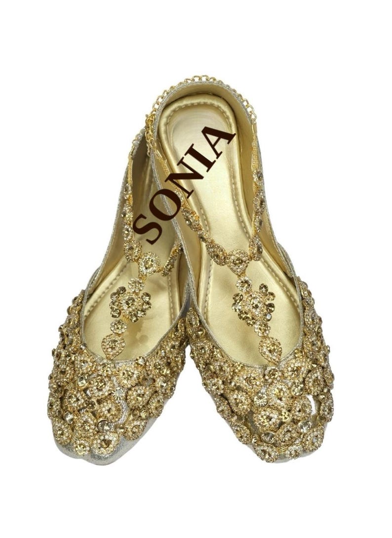 May include: A pair of gold embellished flat shoes with a t-strap design. The shoes are decorated with rhinestones and have a gold chain detail. The word "SONIA" is written on the insole of one shoe.