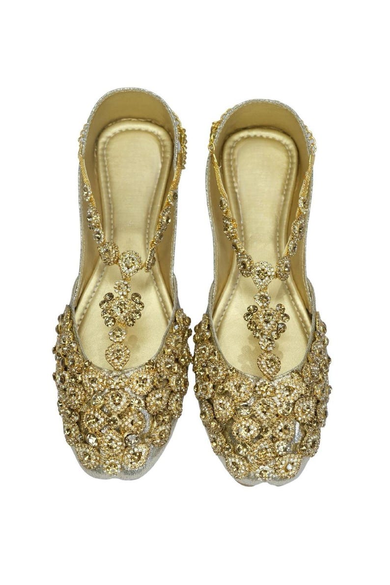 May include: A pair of gold embellished flat shoes with a t-strap design. The shoes are covered in small, sparkling rhinestones.