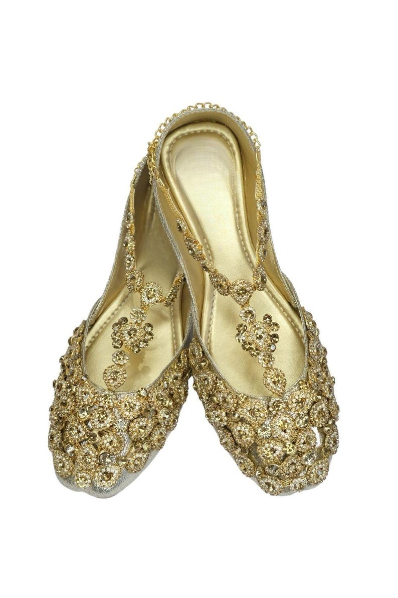 May include: A pair of gold embellished flat shoes with intricate detailing. The shoes have a pointed toe and a delicate ankle strap.