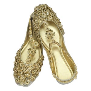 May include: A pair of gold embellished flat shoes with a delicate chain detail. The shoes are adorned with sparkling rhinestones and have a pointed toe.
