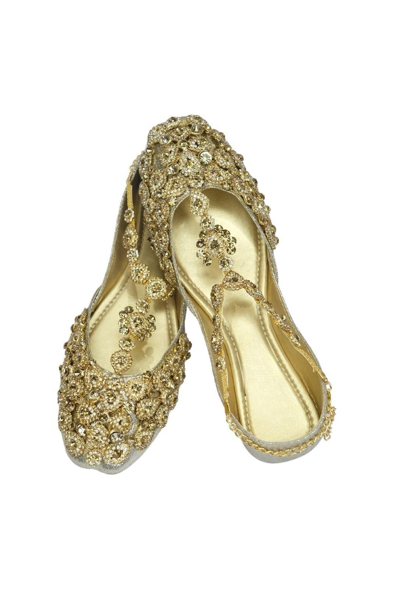 May include: A pair of gold embellished flat shoes with a delicate chain detail. The shoes are adorned with sparkling rhinestones and have a pointed toe.