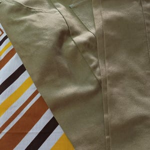 Op de afbeelding: Khaki broek met een klassiek vijf-zakken ontwerp. De broek is gemaakt van een gladde, geweven stof en ligt op een oppervlak met patronen met bruine, gele en witte strepen en bloemvormen.