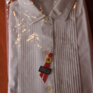 Pode incluir: Uma camisa branca de vestir com frente plissada e colarinho. A camisa está embalada em um saco de plástico transparente e tem uma etiqueta vermelha e amarela que diz "Divissa" e apresenta um logotipo da Ferrari.