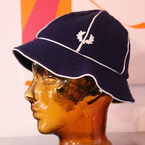 Op de afbeelding: Een navy blauwe bucket hat met een witte rand en een wit geborduurd logo. De hoed zit op een bruin mannequin hoofd.