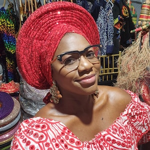 May include: A woman wearing a red sequined head wrap and a white and red embroidered blouse. She is wearing glasses and has a warm smile.