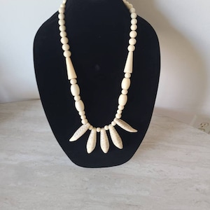 May include: A white beaded necklace with a pendant of five carved bone pieces. The necklace is displayed on a black velvet bust.