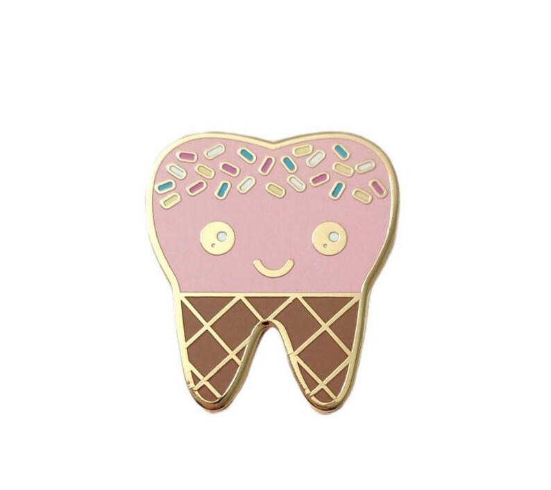 May include: A gold-tone enamel pin shaped like a tooth with a pink ice cream cone on top and brown waffle cone sprinkles. The tooth has a happy face.
