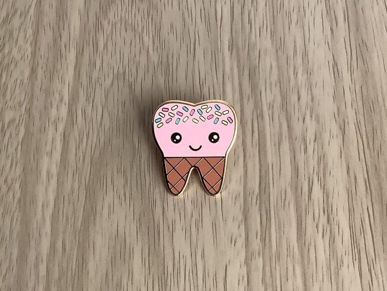 May include: A pink enamel pin shaped like a tooth with a brown cone and colorful sprinkles on top. The tooth has a cute face with eyes and a smile.