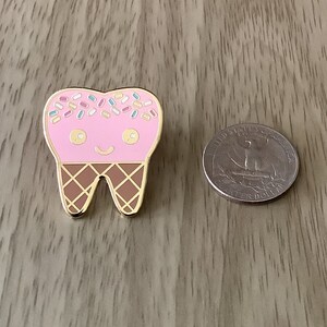 May include: A gold enamel pin shaped like a tooth with a pink ice cream cone on top. The ice cream cone has colorful sprinkles and a happy face.