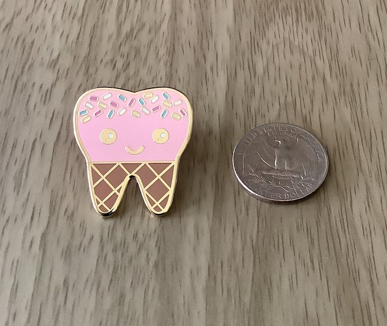 May include: A gold enamel pin shaped like a tooth with a pink ice cream cone on top. The ice cream cone has colorful sprinkles and a happy face.