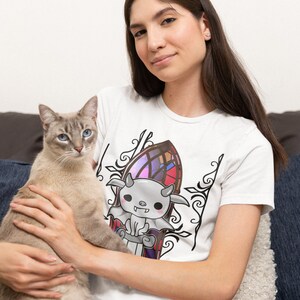 以下が含まれることがあります： 白いTシャツを着た女性が、翼と角を持つ漫画の生き物の白黒のグラフィックを身に着けています。生き物はステンドグラスの窓の前にいます。女性は腕の中に灰色と白の猫を抱えています。