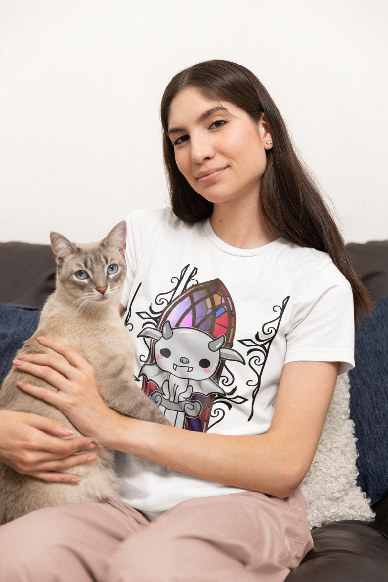 以下が含まれることがあります： 白いTシャツを着た女性が、翼と角を持つ漫画の生き物の白黒のグラフィックを身に着けています。生き物はステンドグラスの窓の前にいます。女性は腕の中に灰色と白の猫を抱えています。