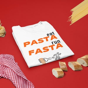 May include: White t-shirt with the text "eat PASTA run FASTA" in orange letters. A black and white illustration of a person running with a fork and pasta on it is below the text. There is a red and white checked cloth, a pile of pasta, and a baguette on a red background.