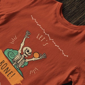 May include: Orange t-shirt with a skeleton surfing on a wave with the text "Let's Bone!" printed on it.