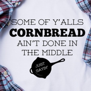 Puede incluir: Camiseta blanca con texto negro que dice "Some of y'alls cornbread ain't done in the middle. Just sayin'" y una silueta negra de una sartén de hierro fundido.