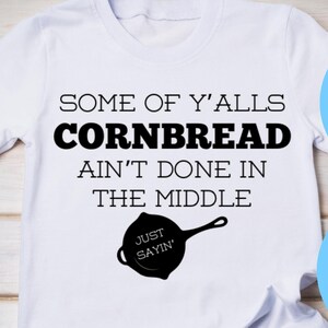 Puede incluir: Camiseta blanca con texto negro que dice "Some of y'alls cornbread ain't done in the middle. Just sayin'" y una silueta negra de una sartén de hierro fundido. La camiseta está sobre una superficie de madera con tela vaquera azul y una cinta morada en el fondo.