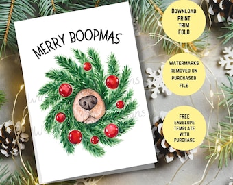 Può includere: Un biglietto di Natale con un'illustrazione ad acquerello di un muso di cane che spunta da una ghirlanda di rami di pino verdi e palline rosse. Il testo "Merry Boopmass" è scritto in nero nella parte superiore della carta.