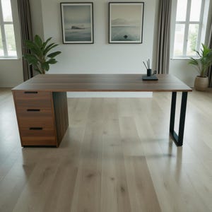 May include: A modern wooden desk with a dark brown tabletop and a three-drawer cabinet on the left side. The desk has black metal legs and is set against a white wall with framed artwork.