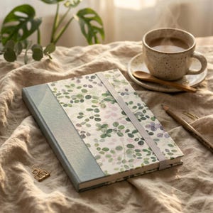 Può includere: Un taccuino rettangolare caratterizzato da un design a copertina divisa con un dorso in tessuto blu-grigio tinta unita e un pannello bianco decorato con un motivo di piccole foglie verdi e viola chiaro, fissato da un elastico grigio chiaro con una graffetta.