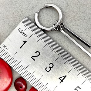 Può includere: Un orecchino a cerchio d'argento con un pendente a forma di barra. L'orecchino viene misurato con un righello, mostrando che il pendente è lungo circa 9 cm.