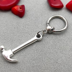 May include: A silver hammer charm attached to a silver hoop earring. The earring is a simple hoop with a ball closure.