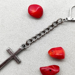 Könnte beinhalten: Ein silberner Kettenohrring mit einem Kreuzanhänger. Der Ohrring hat einen Hakenverschluss. Das Kreuz ist schwarz und hat ein schlichtes Design. Der Ohrring ist von roten Korallensteinen umgeben.