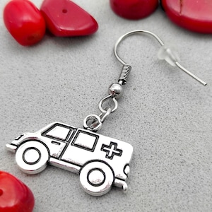 May include: Silver earring featuring a small, detailed charm of a silver ambulance with a red cross. The earring has a silver hook and a clear plastic back.