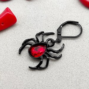 Könnte beinhalten: Schwarzer Spinnenohrring mit einem roten Edelstein. Der Ohrring hat einen Hakenverschluss.