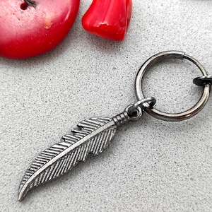 May include: A silver feather charm attached to a silver hoop earring. The earring is a clip-on style.