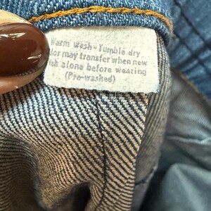 May include: A close-up of a white care label sewn into a pair of blue jeans. The label reads "Warm wash Tumble dry Color may transfer when new Wash alone before wearing (Pre-washed)"