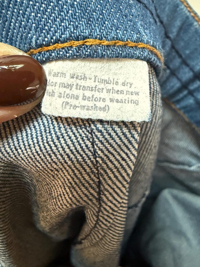 May include: A close-up of a white care label sewn into a pair of blue jeans. The label reads "Warm wash Tumble dry Color may transfer when new Wash alone before wearing (Pre-washed)"