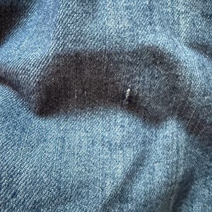 May include: Close-up of a blue denim fabric with a textured weave. The fabric is folded and wrinkled, showing the natural texture of the material.