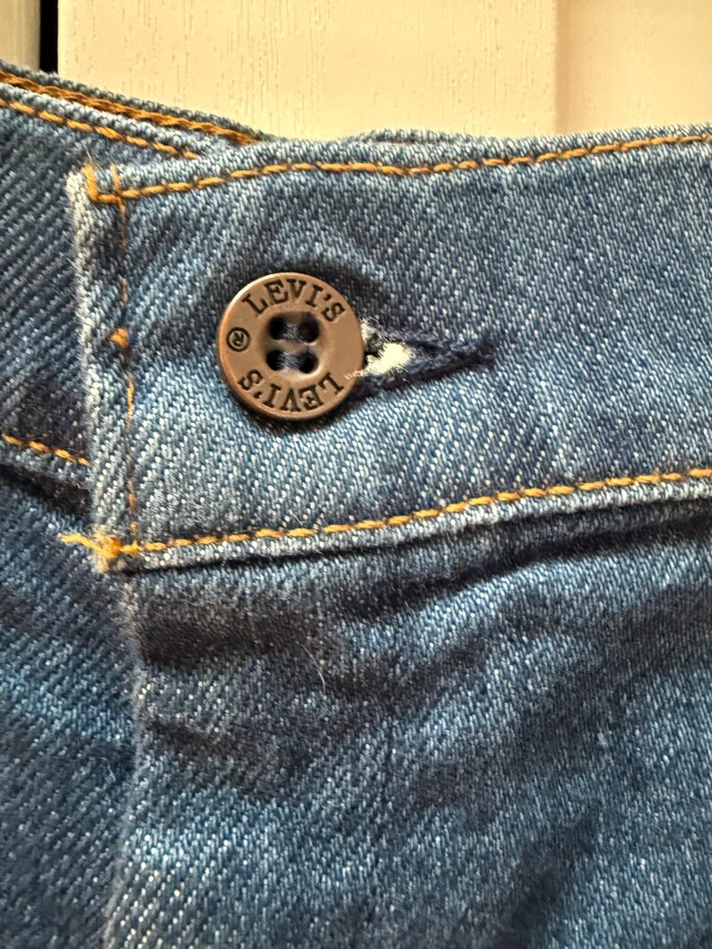 May include: Close-up of a blue denim jean with a brown button that says "LEVI'S" in a circular design.