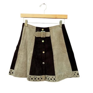 Peut inclure: Jupe trapèze en daim marron, beige et noir. La jupe est ornée d'une boucle, de boutons décoratifs et d'un ourlet découpé au laser. La jupe est suspendue à un cintre en bois.