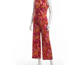 May include: A sleeveless jumpsuit in a vibrant, geometric floral pattern. The jumpsuit features a mix of red, pink, and orange hues. The wide-leg design is displayed on a white mannequin, showcasing the vintage style of the garment.