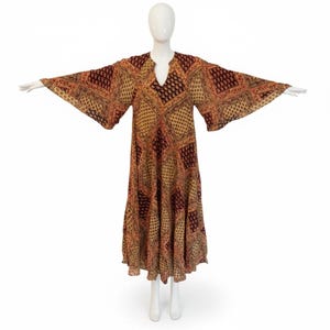 Puede incluir: Un vestido largo y fluido con mangas anchas, exhibido en un maniquí. El vestido presenta un cuello en V y un estampado de retazos en tonos marrón, naranja y amarillo. La tela parece ligera y aireada, adecuada para climas cálidos.